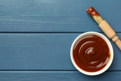 Photo of Tasty barbeque sauce in bowl and brush on blue wooden table, top view. Space for text