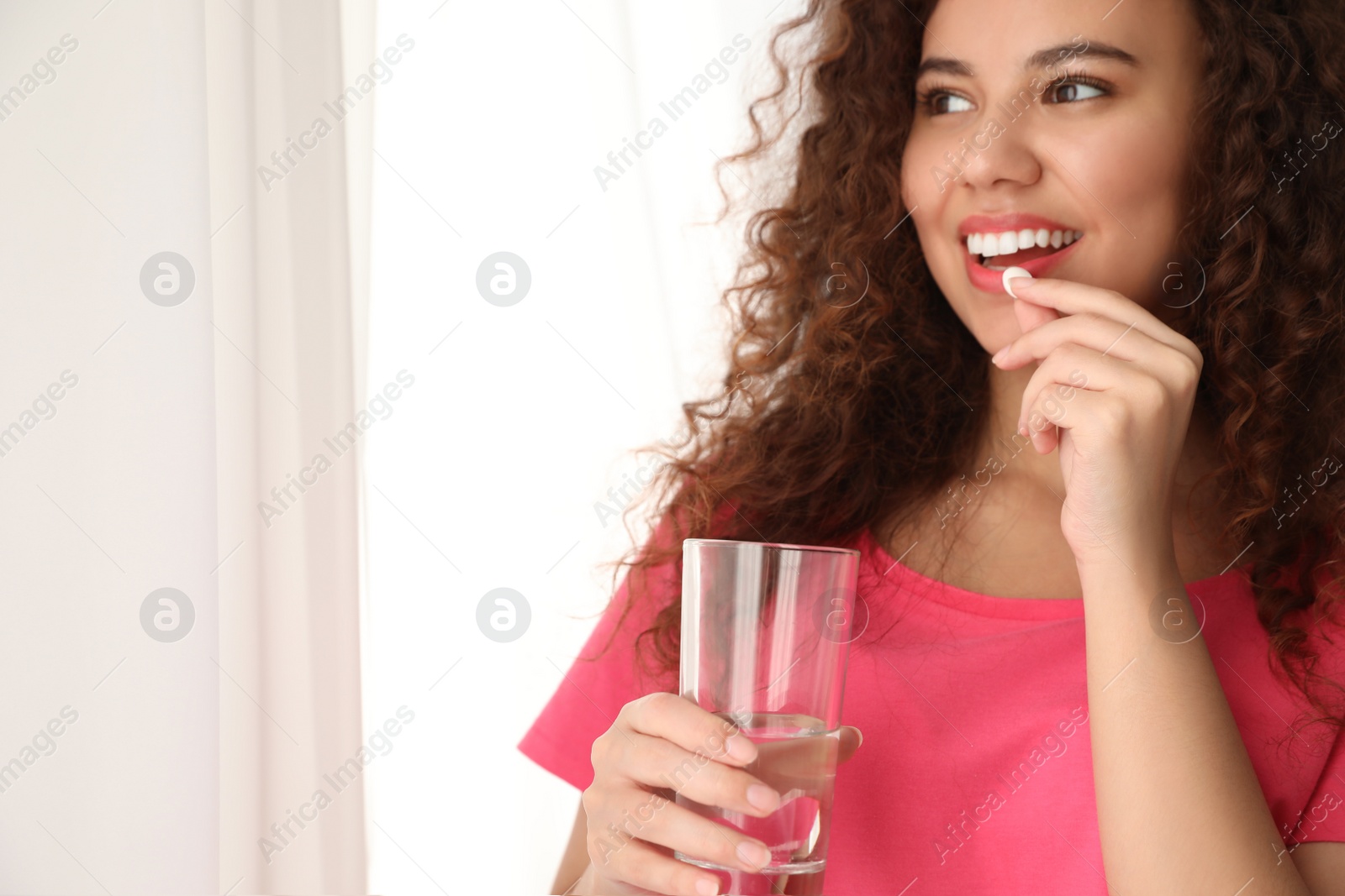 Photo of African-American woman with glass of water taking vitamin pill on light background. Space for text