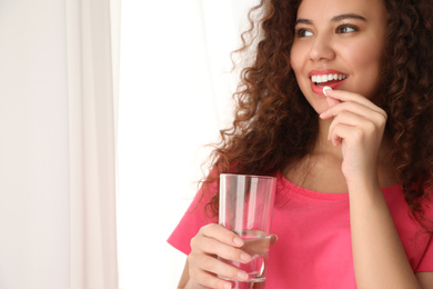 African-American woman with glass of water taking vitamin pill on light background. Space for text