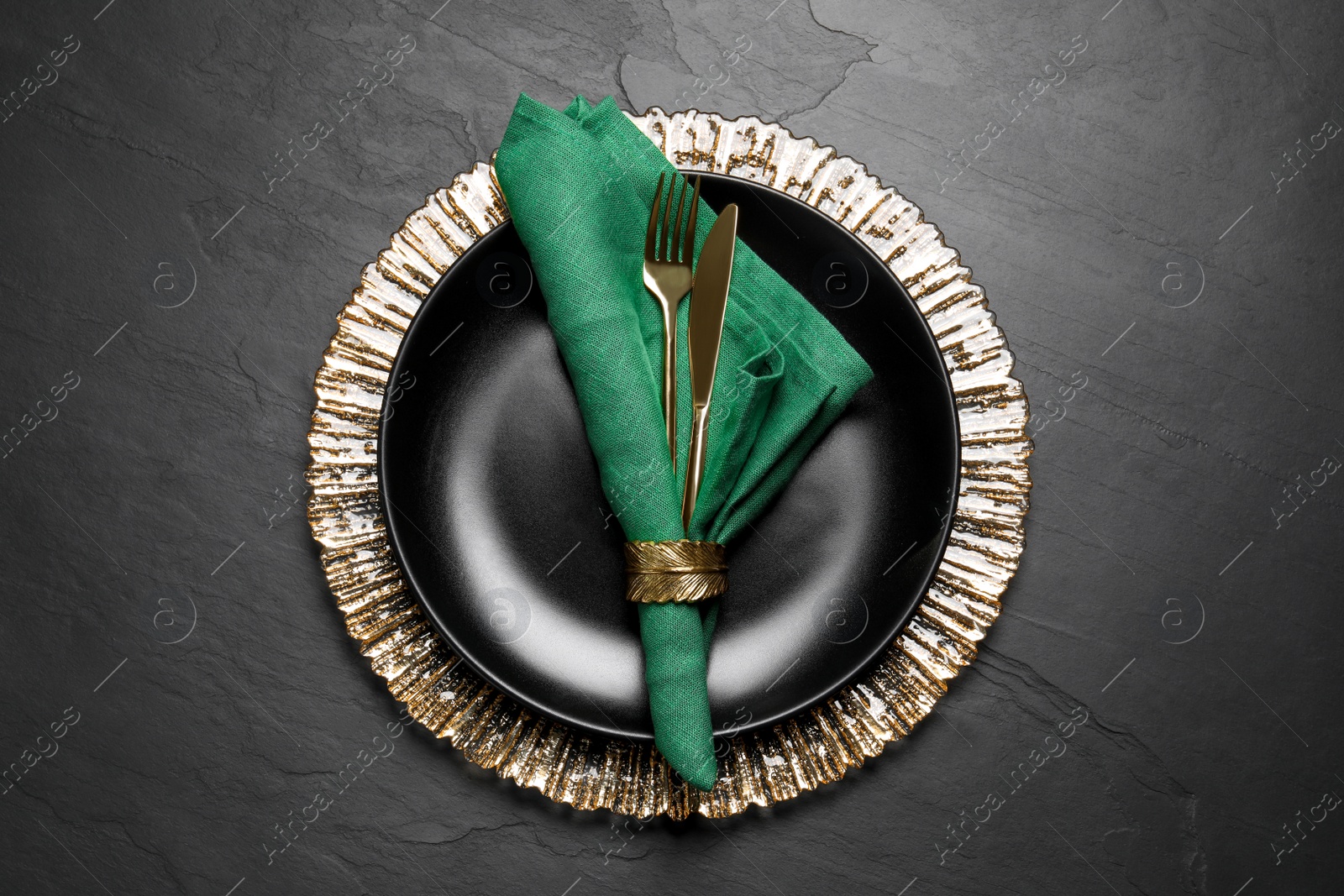 Photo of Plate with green fabric napkin, decorative ring and cutlery on black table, top view