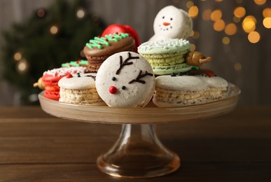 Photo of Stand with beautifully decorated Christmas macarons on wooden table against blurred festive lights