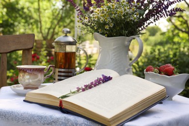 Open book, tea, ripe strawberries and bouquet of beautiful wildflowers on table in garden