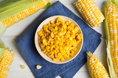 Tasty sweet corn kernels in bowl and fresh cobs on white wooden table, flat lay