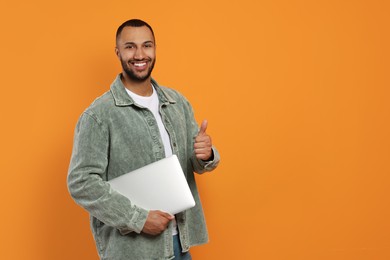 Photo of Smiling young man with laptop showing thumbs up on orange background, space for text
