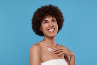 Beautiful young woman applying cream onto body on light blue background