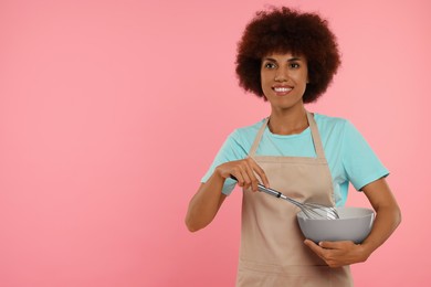 Photo of Happy young woman in apron with whisk and bowl on pink background. Space for text