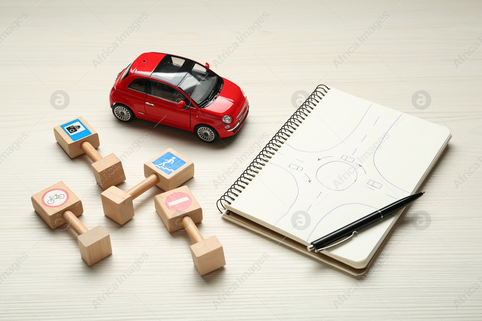 Photo of Different miniature road signs, car and notebook with sketch of roundabout on white wooden background. Driving school