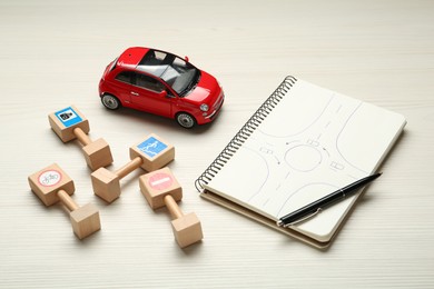 Photo of Different miniature road signs, car and notebook with sketch of roundabout on white wooden background. Driving school