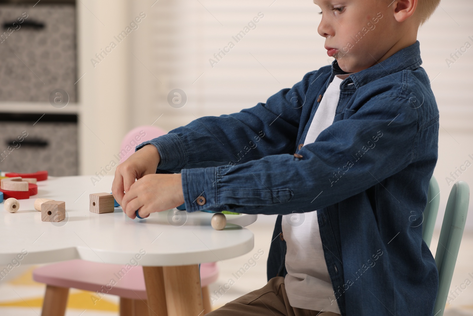 Photo of Little boy playing with wooden pieces and string for threading activity at white table indoors, closeup. Child's toy