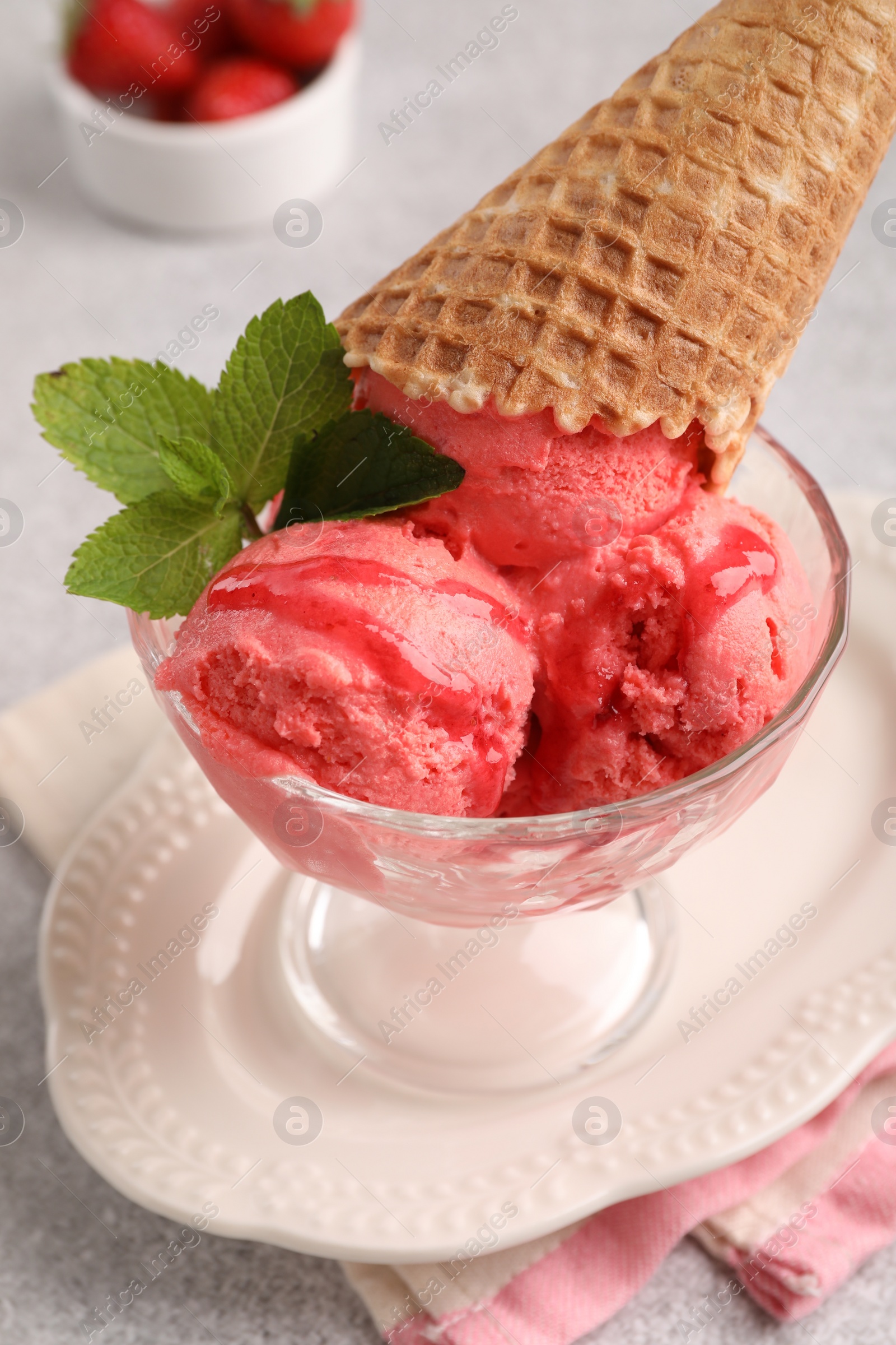 Photo of Delicious scoops of strawberry ice cream with mint and wafer cone in glass dessert bowl served on grey table, closeup