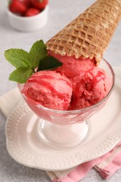 Photo of Delicious scoops of strawberry ice cream with mint and wafer cone in glass dessert bowl served on grey table, closeup