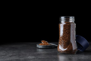 Photo of Glass jar and spoon of instant coffee on grey table against black background. Space for text