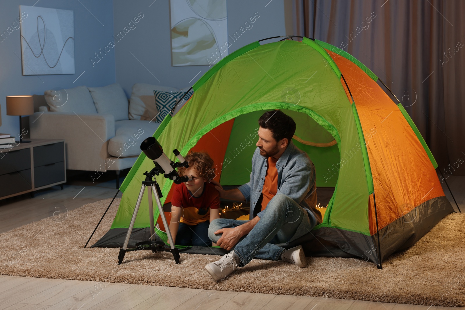 Photo of Father and his son using telescope to look at stars while sitting in camping tent indoors