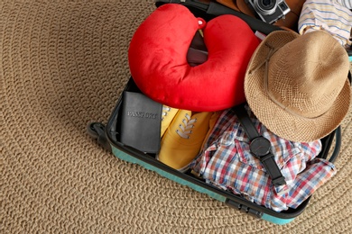 Photo of Opened suitcase with travel pillow and clothes on wicker rug