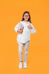 Happy schoolgirl with books showing thumb up on orange background
