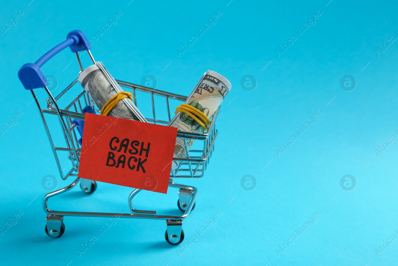 Photo of Rolled dollar banknotes in shopping cart on light blue background, space for text. Cashback concept