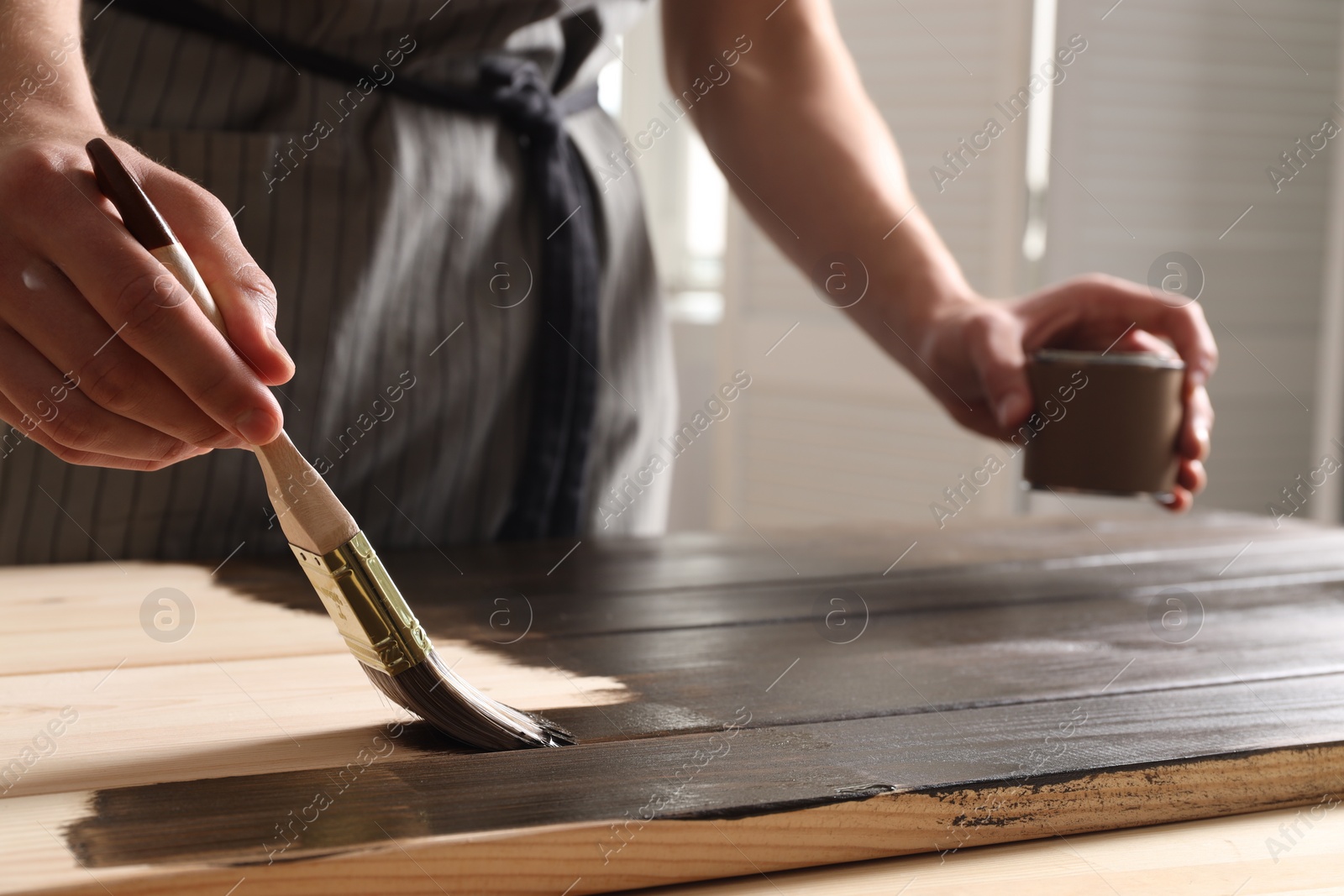 Photo of Man with brush and can applying wood stain onto wooden surface indoors, closeup