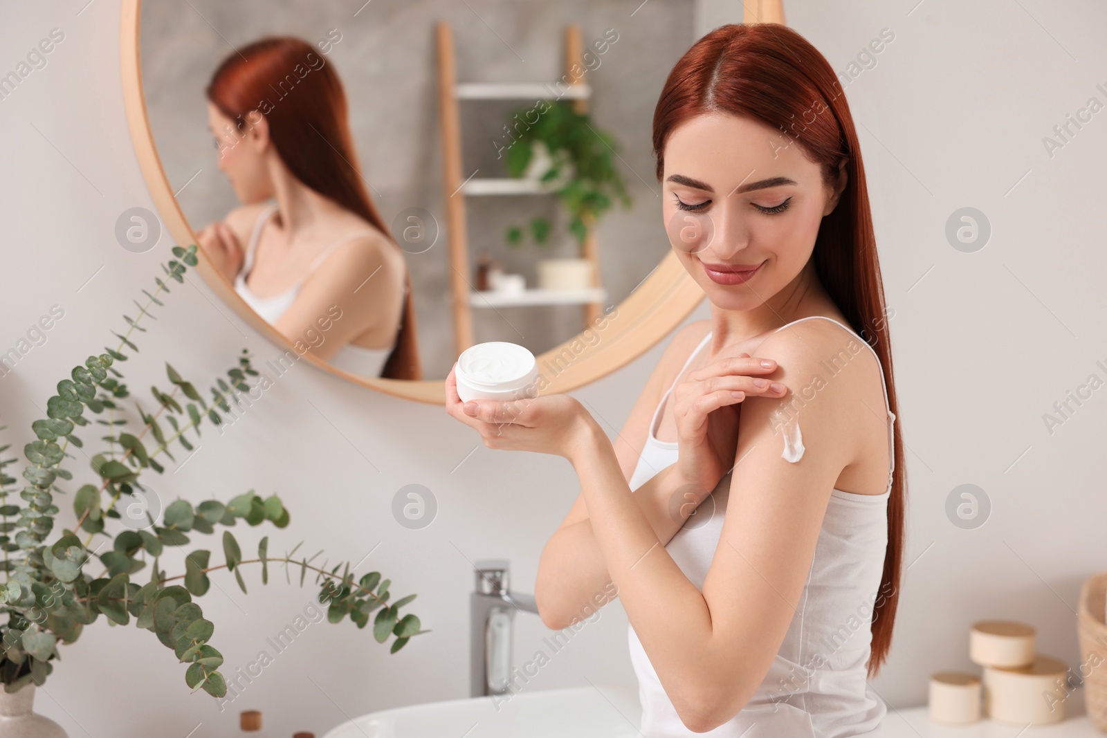 Photo of Beautiful young woman applying body cream onto shoulder in bathroom, space for text