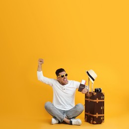 Emotional male tourist holding passport with ticket near suitcase on yellow background