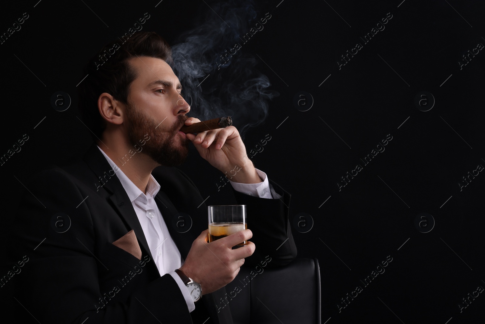 Photo of Handsome man in elegant suit with glass of whiskey smoking cigar on black background. Space for text