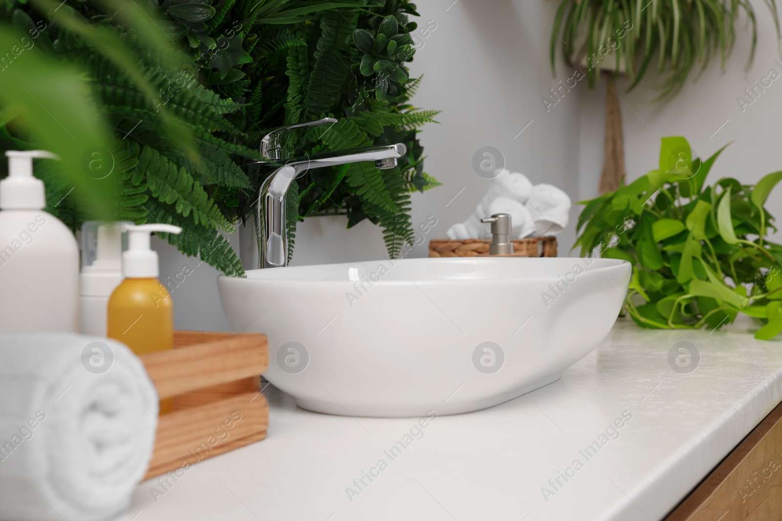 Photo of Green artificial plants, vessel sink and different personal care products in bathroom