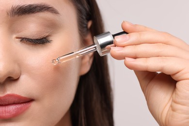 Young woman applying essential oil onto face on light grey background, closeup