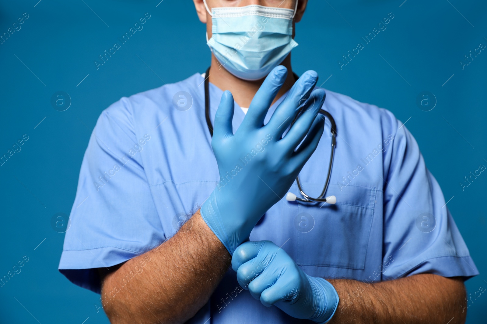 Photo of Doctor in protective mask putting on medical gloves against blue background, closeup