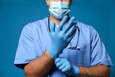 Doctor in protective mask putting on medical gloves against blue background, closeup