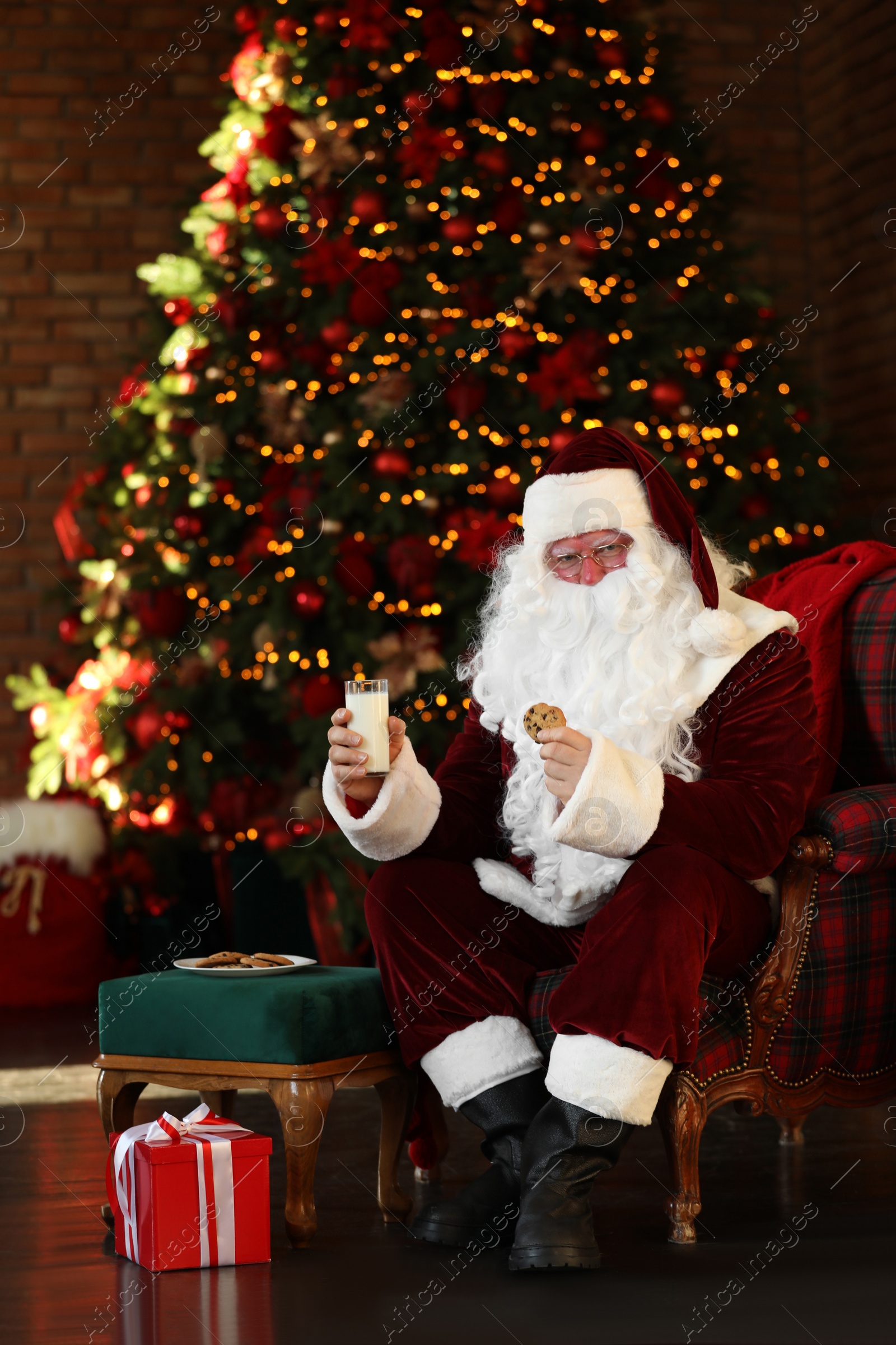 Photo of Santa Claus with milk and cookie near Christmas tree indoors