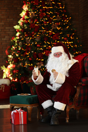 Photo of Santa Claus with milk and cookie near Christmas tree indoors