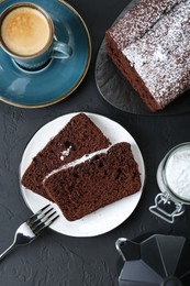 Tasty chocolate sponge cake with powdered sugar and coffee on black textured table, flat lay