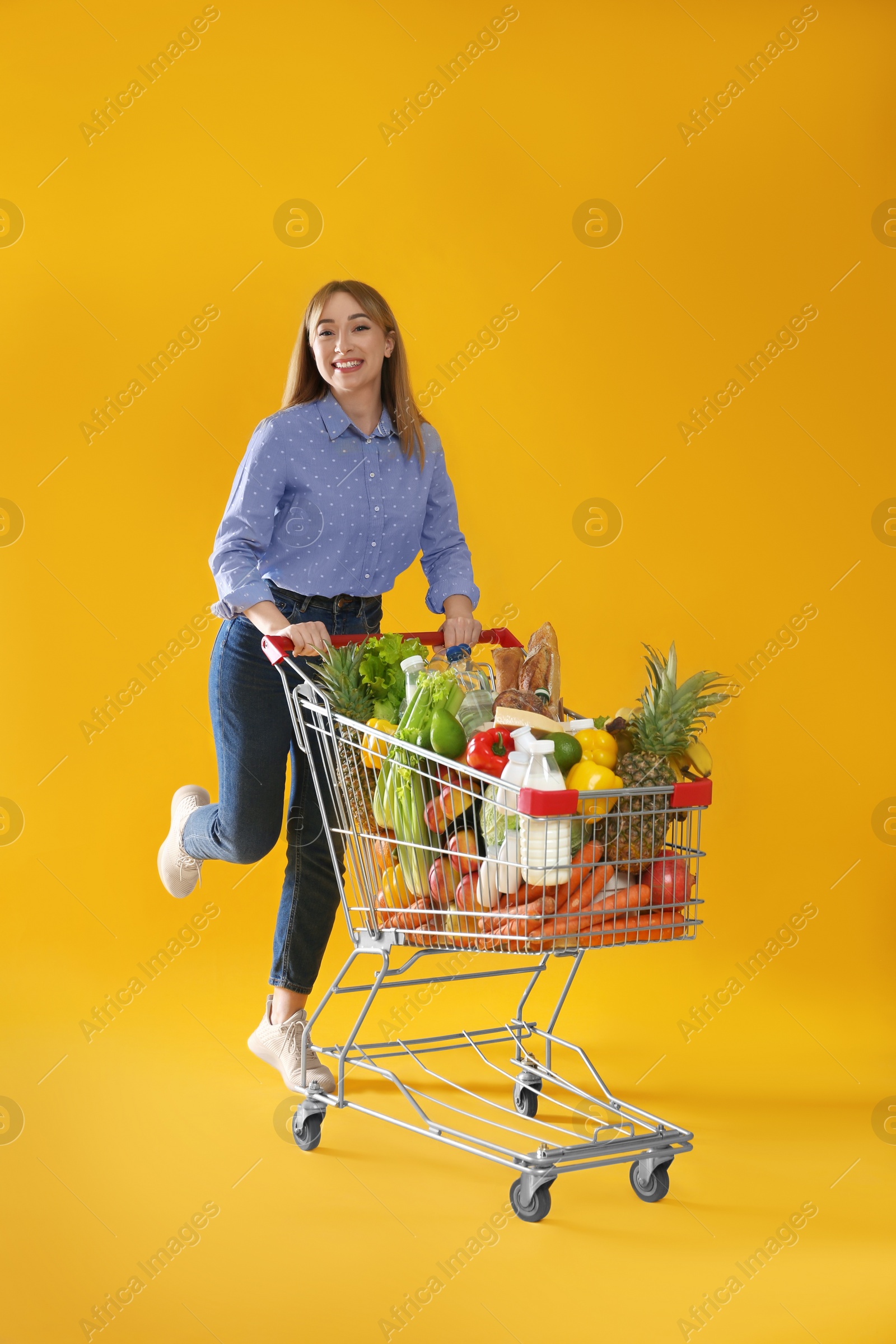 Photo of Young woman with shopping cart full of groceries on yellow background