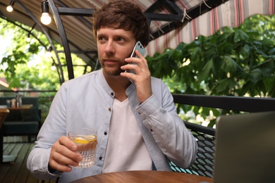 Photo of Handsome man with glass of water talking on phone at table in outdoor cafe