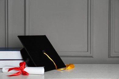 Photo of Graduation hat, books and diploma on white table near grey wall, space for text