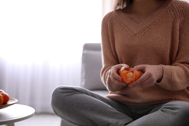 Photo of Young woman peeling fresh tangerine on sofa at home, closeup