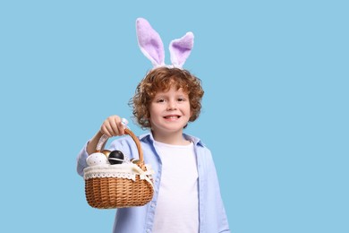 Photo of Portrait of happy boy in cute bunny ears headband holding wicker basket with Easter eggs on light blue background