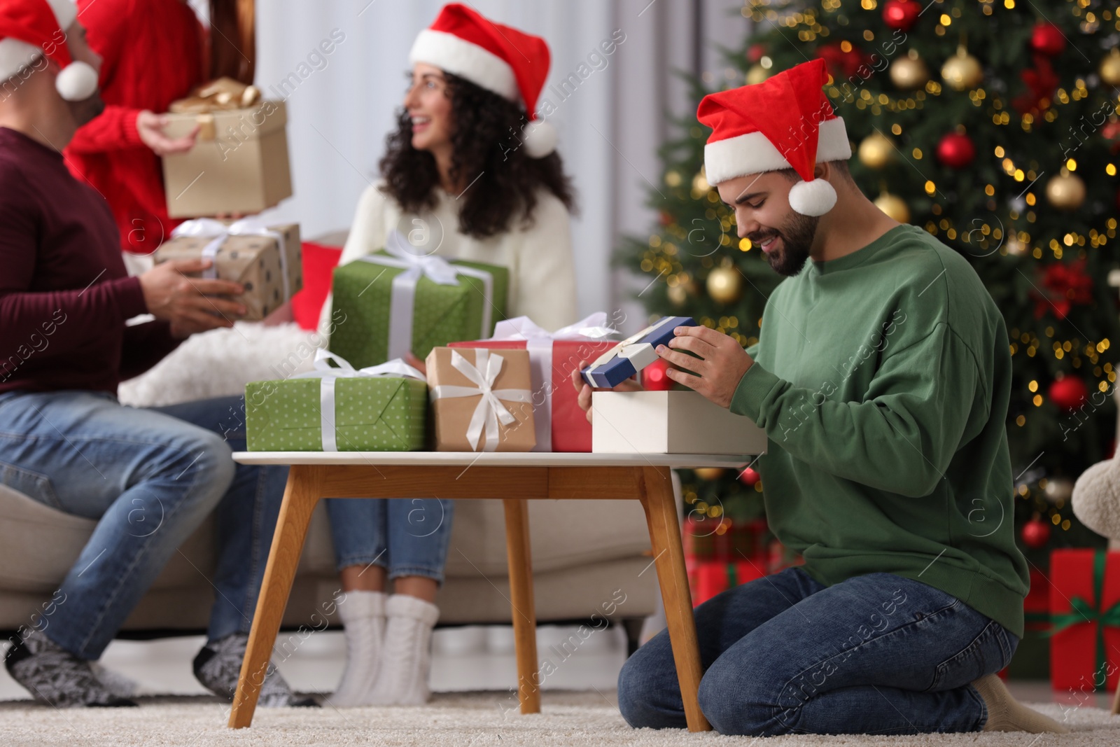 Photo of Christmas celebration in circle of friends. Happy young man opening gift at home, selective focus