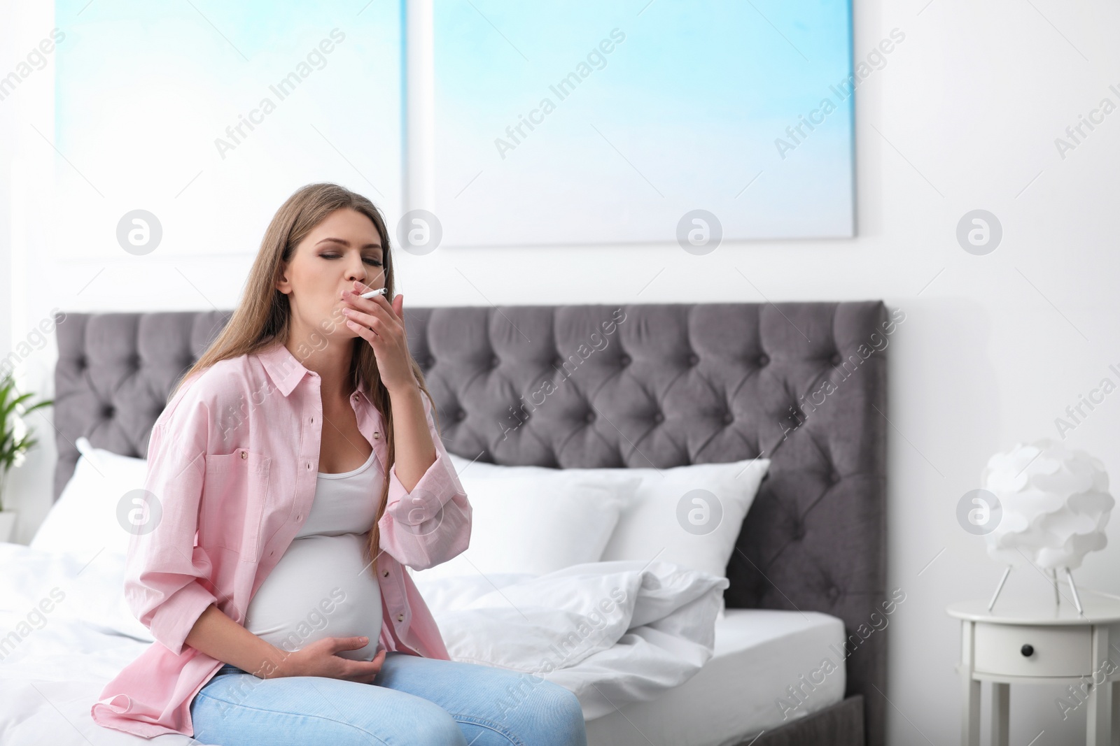 Photo of Young pregnant woman smoking cigarette in bedroom. Harm to unborn baby