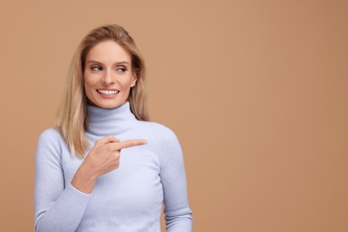 Photo of Portrait of smiling middle aged woman with blonde hair pointing at something on beige background. Space for text