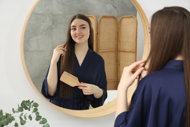 Beautiful woman in blue robe brushing hair near mirror indoors