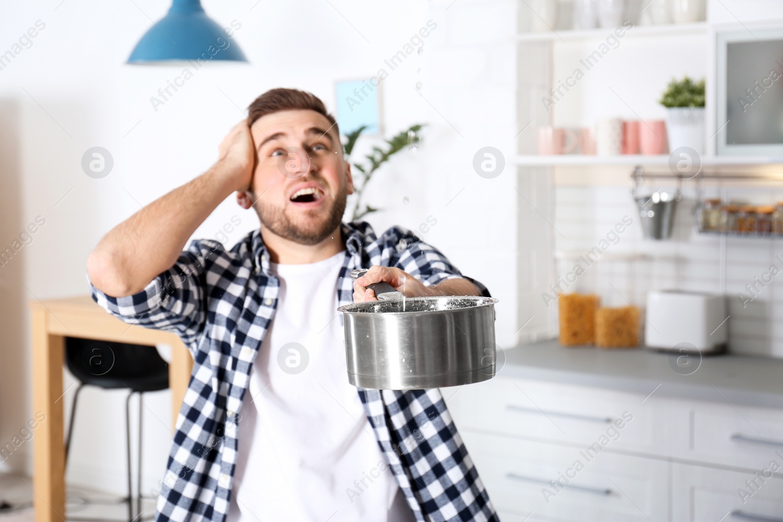 Photo of Emotional young man holding saucepan under water leakage from ceiling in kitchen, space for text. Plumber service