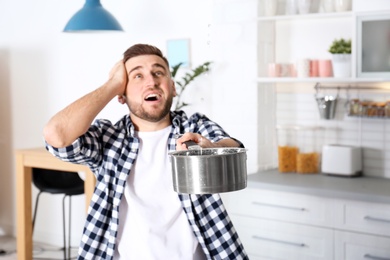 Photo of Emotional young man holding saucepan under water leakage from ceiling in kitchen, space for text. Plumber service