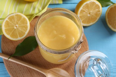 Photo of Delicious lemon curd in glass jar, fresh citrus fruits, spoon and green leaf on light blue wooden table, closeup