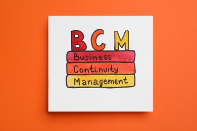 Paper with BCM abbreviation and its interpretation (Business Continuity Management) on orange background, top view
