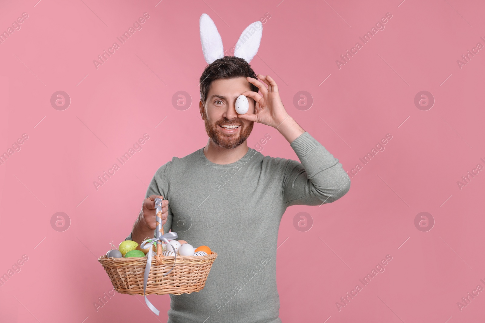 Photo of Happy man in cute bunny ears headband covering eye with Easter egg and holding wicker basket on pink background