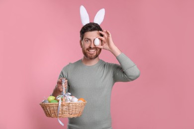 Happy man in cute bunny ears headband covering eye with Easter egg and holding wicker basket on pink background