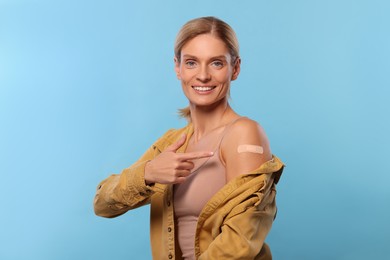 Smiling woman pointing at adhesive bandage on arm after vaccination on light blue background