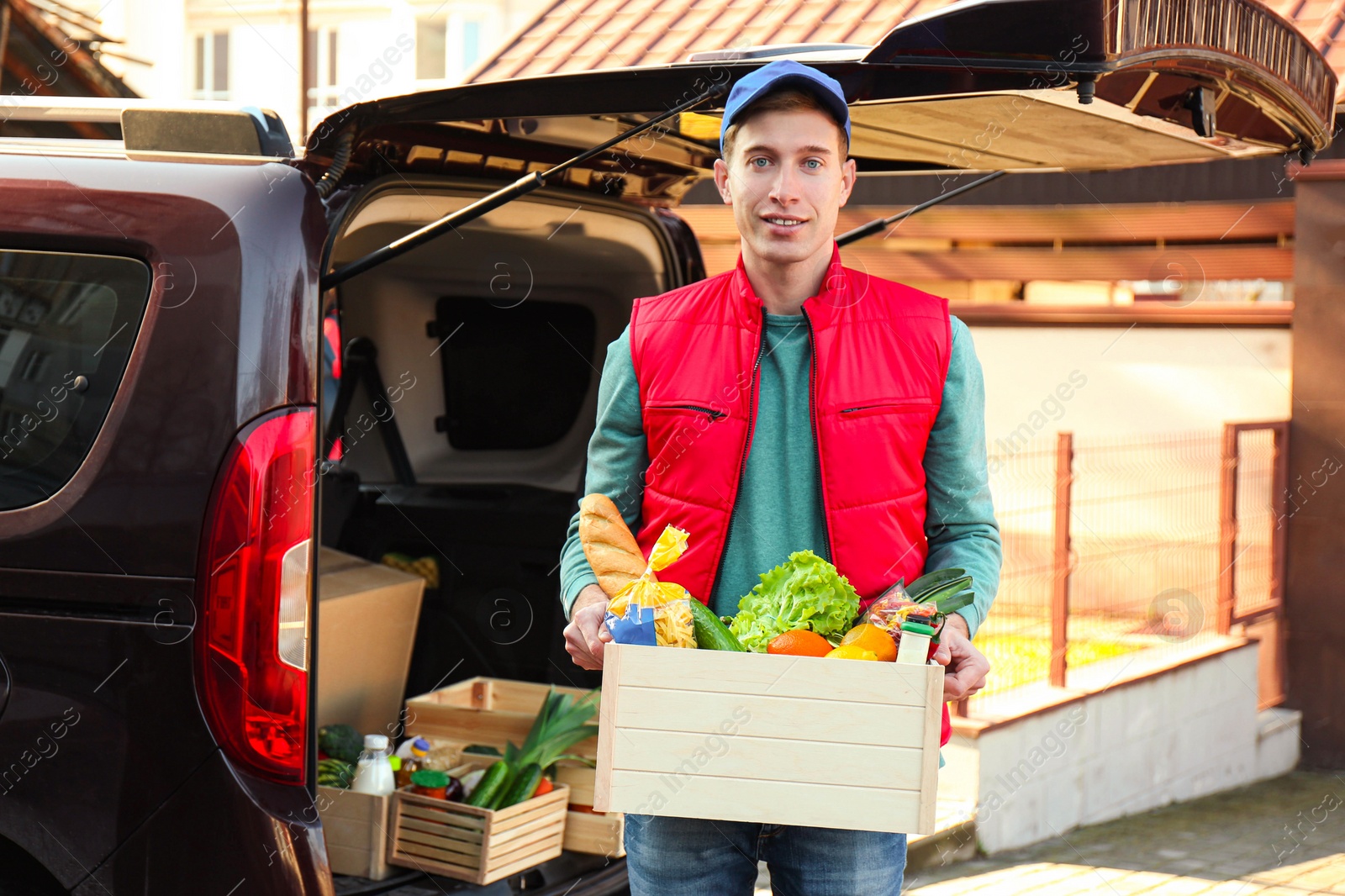 Photo of Courier holding crate with products near car outdoors. Food delivery service