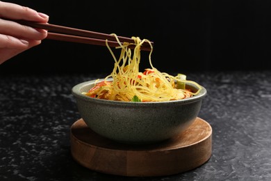 Photo of Stir-fry. Woman using chopsticks for eating tasty noodles with meat at dark textured table, closeup