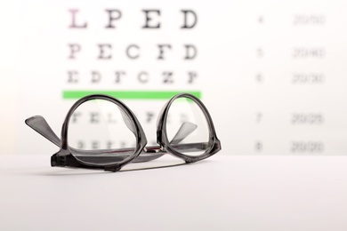 Photo of Glasses with corrective lenses on table against eye chart